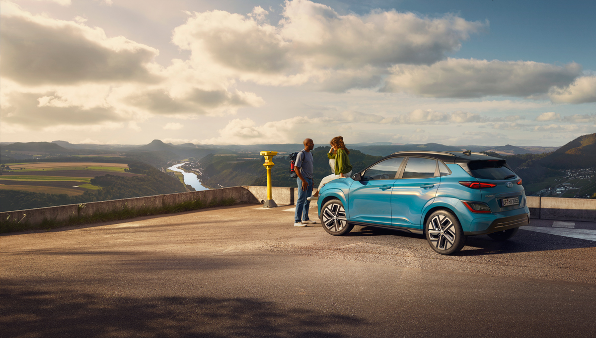 Mladá dvojice sedí na kapotě SUV Hyundai KONA Electric zaparkovaného na místě s výhledem do malebné krajiny.